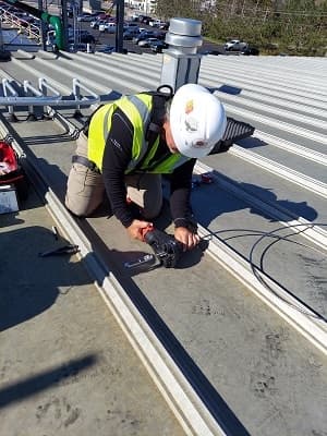Technician load-testing a structural roof anchor, kneeling on a metal roof in hi-vis and a hard hat